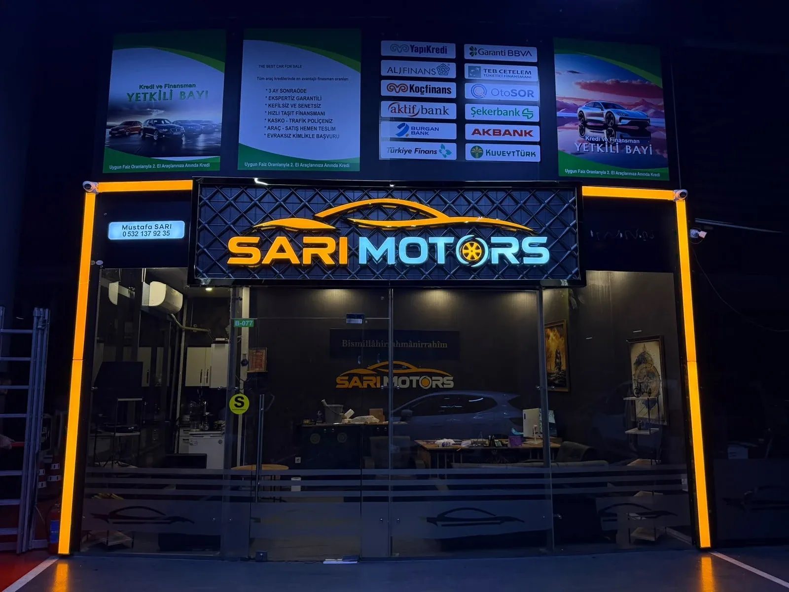 Sarı Motors oto galerisinin kapalı galeri içindeki cephesi: baklava dokulu koyu zemin panosu üzerinde turuncu araç silüeti, ışıklı SARI ve MOTORS yazısı, O harfinin yerinde turuncu jant; vitrinin iki yanında dikey turuncu ışık kolonları