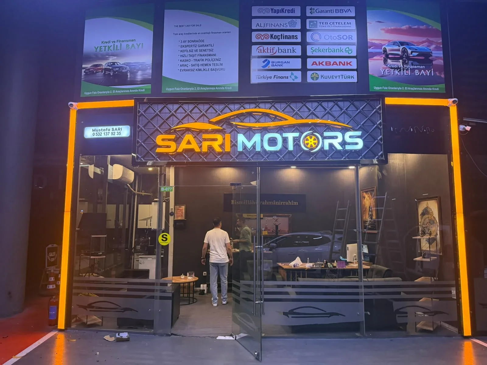 Sarı Motors oto galerisinin kapalı galeri içindeki cephesi: baklava dokulu koyu zemin panosu üzerinde turuncu araç silüeti, ışıklı SARI ve MOTORS yazısı, O harfinin yerinde turuncu jant; vitrinin iki yanında dikey turuncu ışık kolonları — Işıklı kutu harf detayı