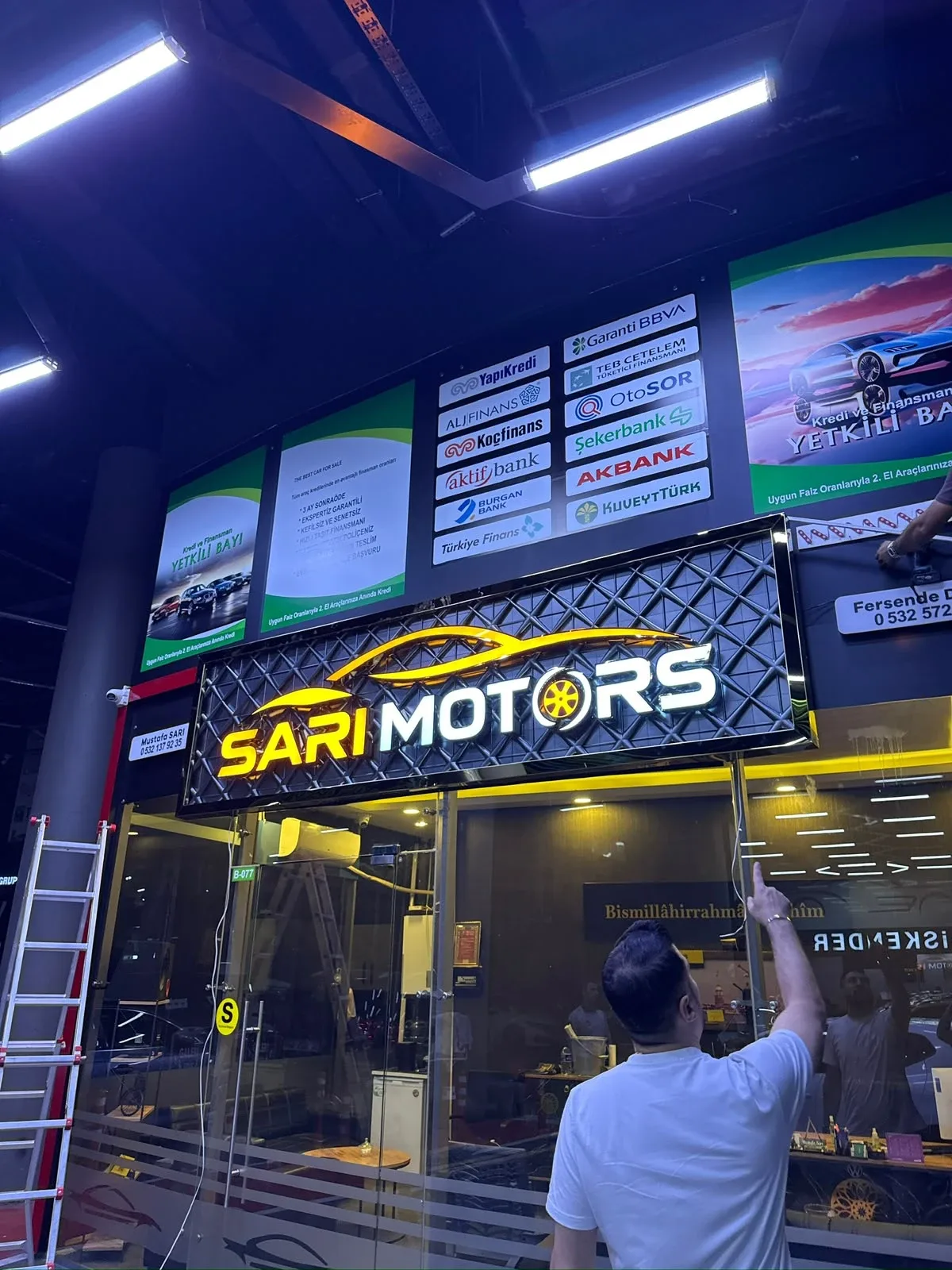 Sarı Motors oto galerisinin kapalı galeri içindeki cephesi: baklava dokulu koyu zemin panosu üzerinde turuncu araç silüeti, ışıklı SARI ve MOTORS yazısı, O harfinin yerinde turuncu jant; vitrinin iki yanında dikey turuncu ışık kolonları