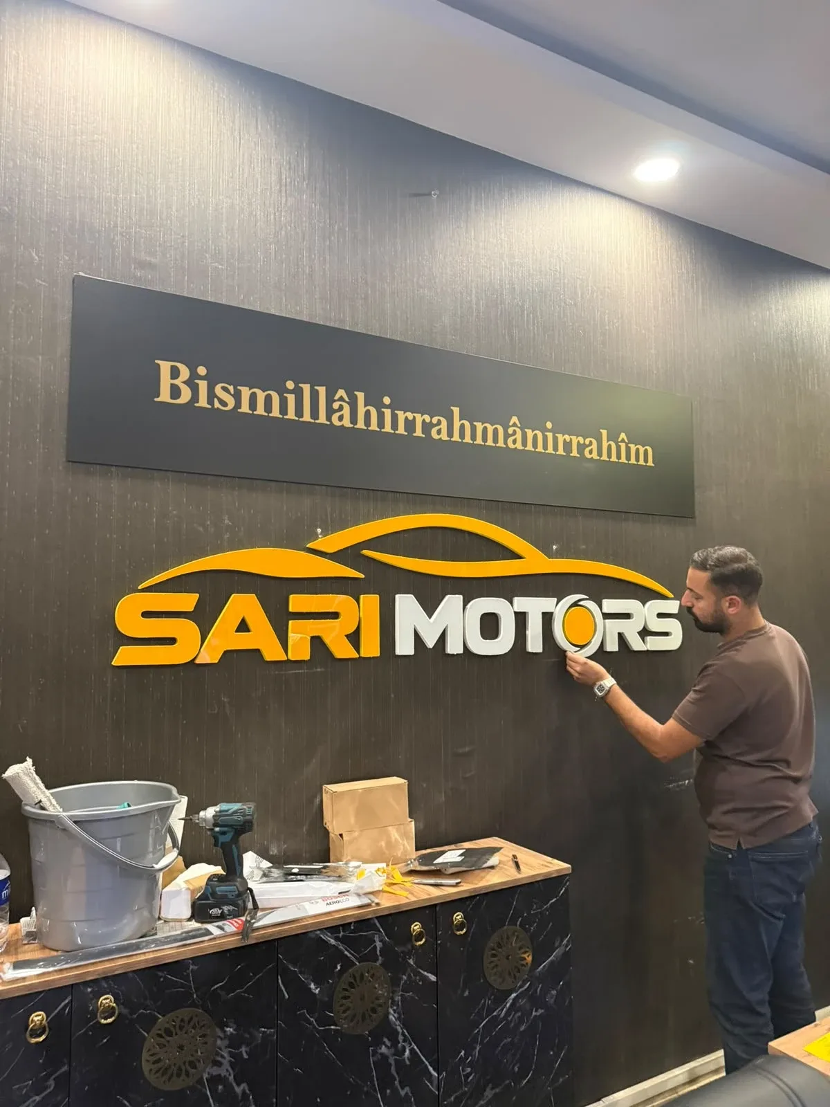 Sarı Motors oto galerisinin kapalı galeri içindeki cephesi: baklava dokulu koyu zemin panosu üzerinde turuncu araç silüeti, ışıklı SARI ve MOTORS yazısı, O harfinin yerinde turuncu jant; vitrinin iki yanında dikey turuncu ışık kolonları — İç mekân kabartma logo yakın plan