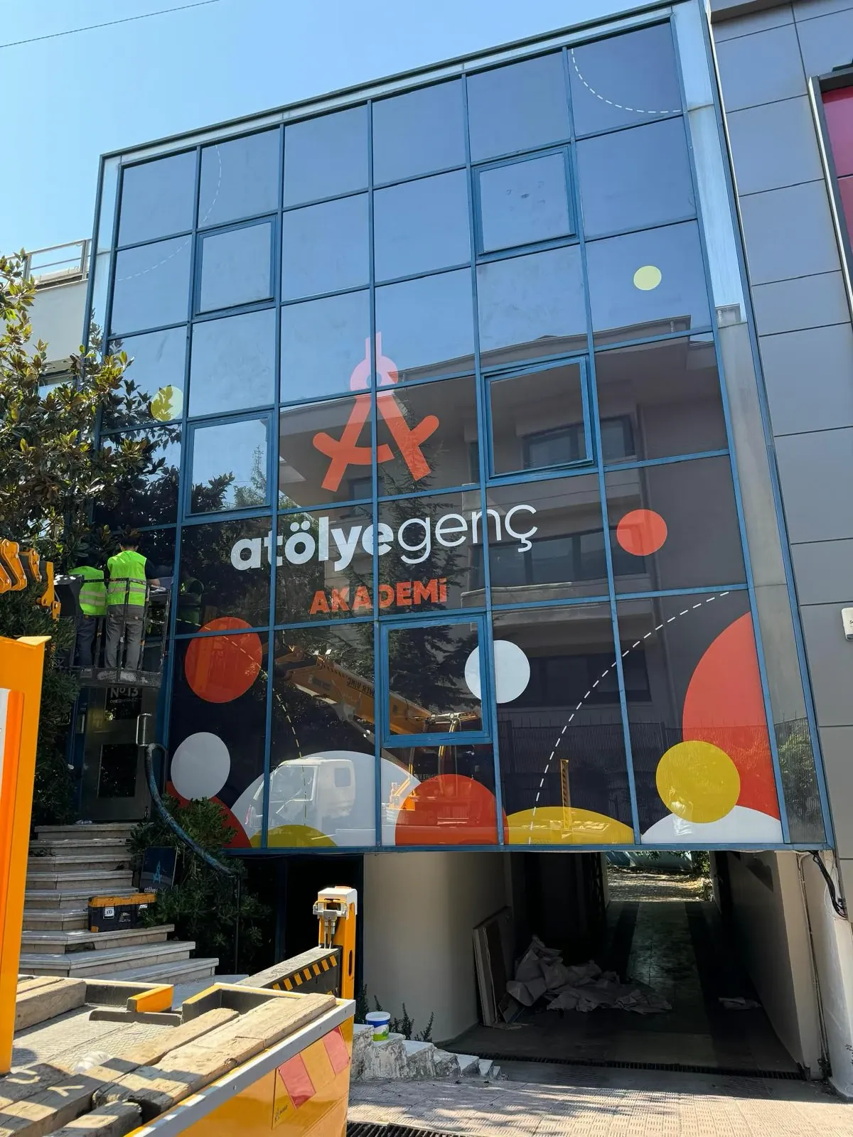 Atölye Genç Akademi binasının mavi profilli cam cephesinde baskılı giydirme: turuncu pergel biçimli logo, atölyegenç AKADEMİ yazısı, turuncu, sarı ve beyaz daireler ile kesik çizgili yaylar; solda vinç sepetinde uygulama yapan ekip