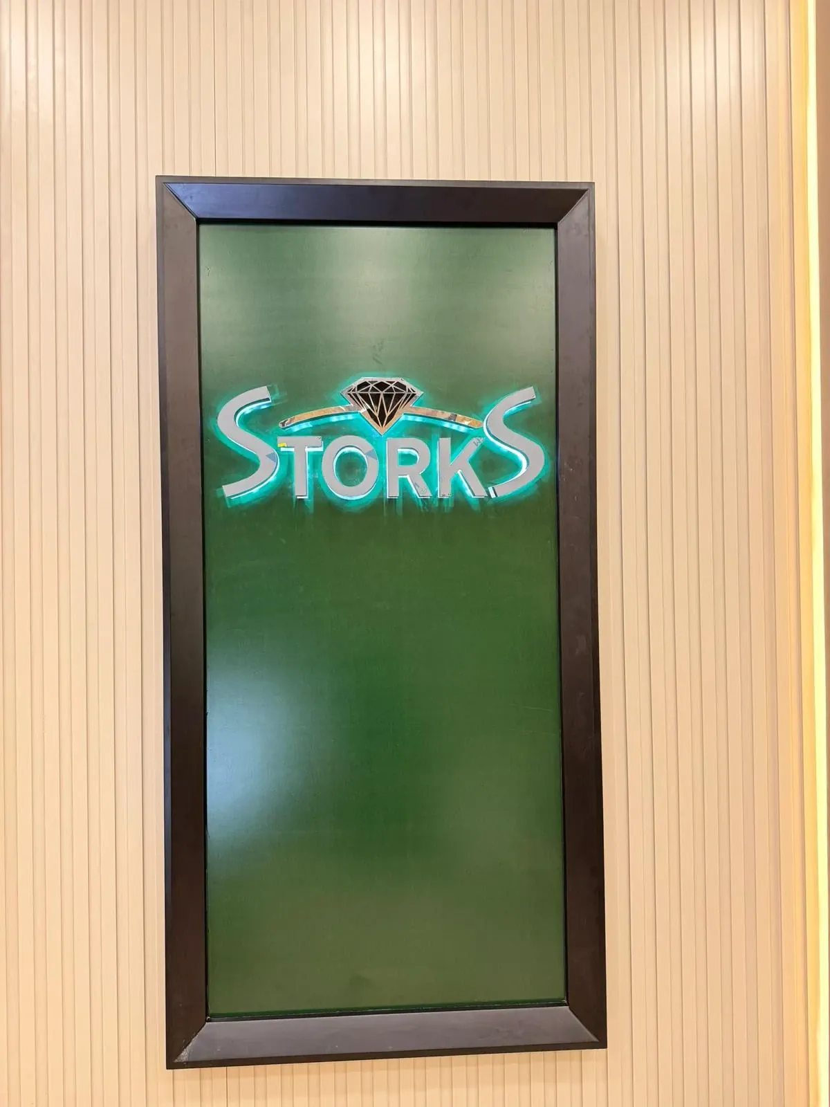 Storks iç mekân logo panosu: dikey çıtalı krem duvar üzerinde koyu çerçeveli yeşil pano, ayna parlaklığında paslanmaz STORKS harfleri ve üstünde siyah pırlanta simgesi; harflerin arkasından turkuaz LED ışığı panoya yansıyor