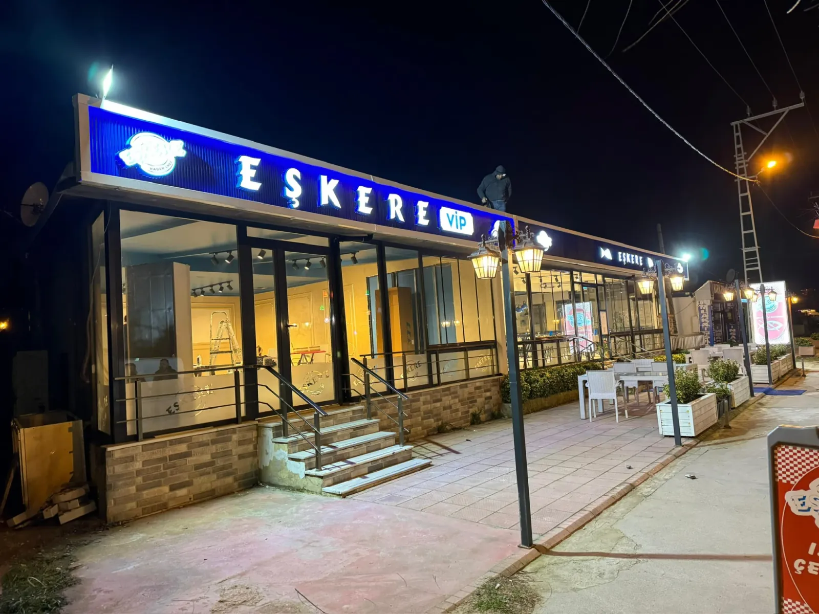 Eşkere restoranının cephesinde gece: mavi ışıklı zemin pano üzerinde beyaz EŞKERE harfleri, beyaz rozet içinde VIP yazısı ve solda yuvarlak logo; panonun üzerinde son ayarı yapan ekipten biri, önde aydınlatılmış teras ve fenerli direkler