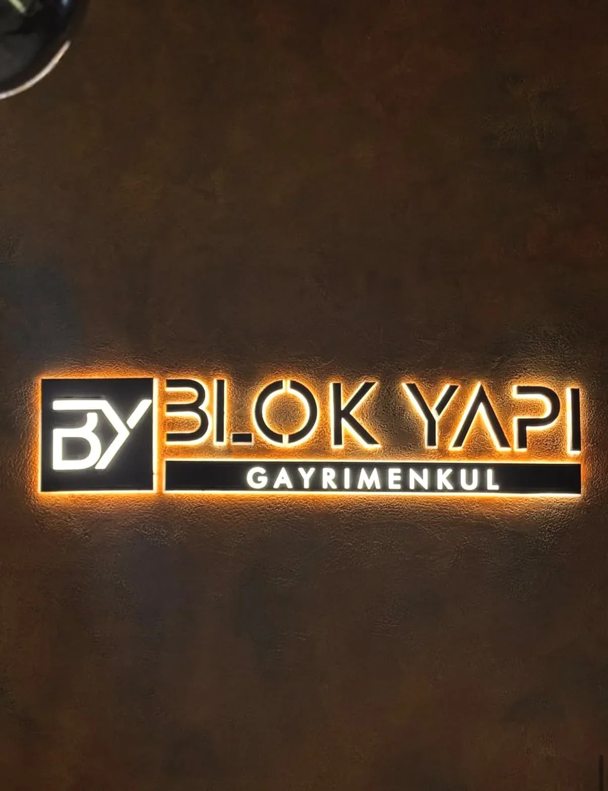 Koyu kahverengi dokulu duvar üzerinde sıcak turuncu halo ışıkla arkadan aydınlatılan siyah yüzeyli kutu harfler ve logo bloğu
